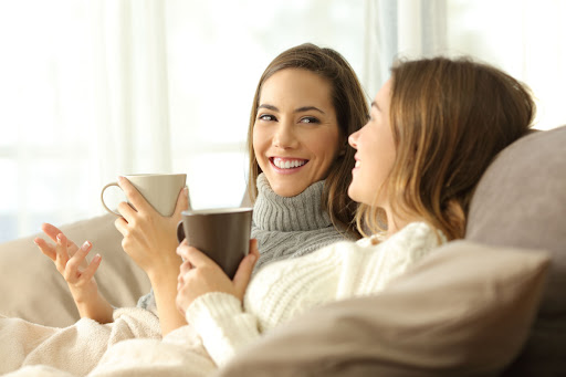 Two women wearing sweaters sitting on a couch with cups of coffee.