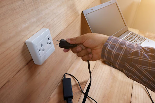 A close-up of a hand plugging a laptop charger into an outlet.