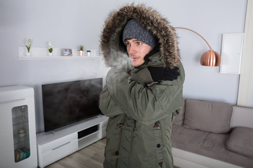 A shivering man wearing a coat in a cold house.