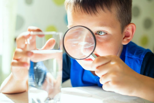 A boy inspecting a glass of water with a magnifying glass.