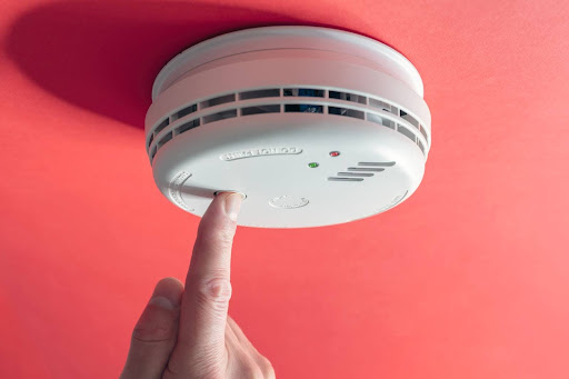 A close-up of a finger pressing a button on a smoke detector on a ceiling.